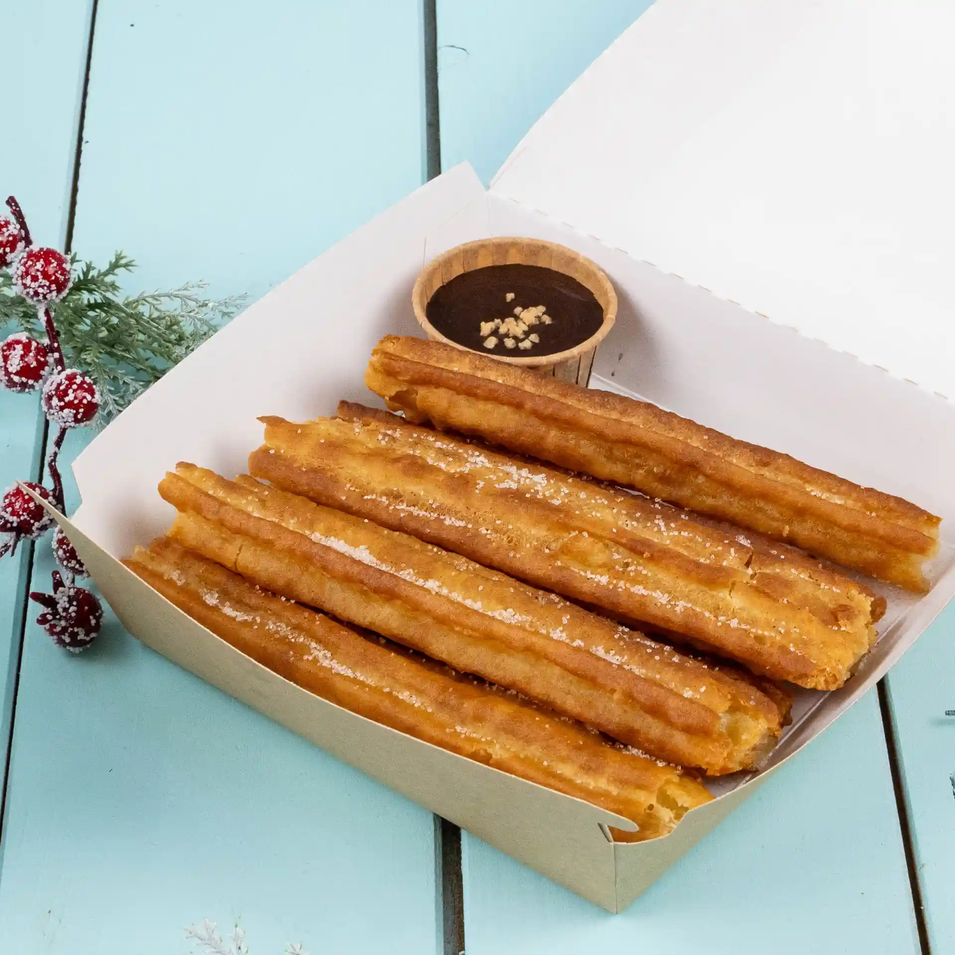 Churros in een papieren doos met een klein bruin kopje chocoladesaus op een blauwe ondergrond.