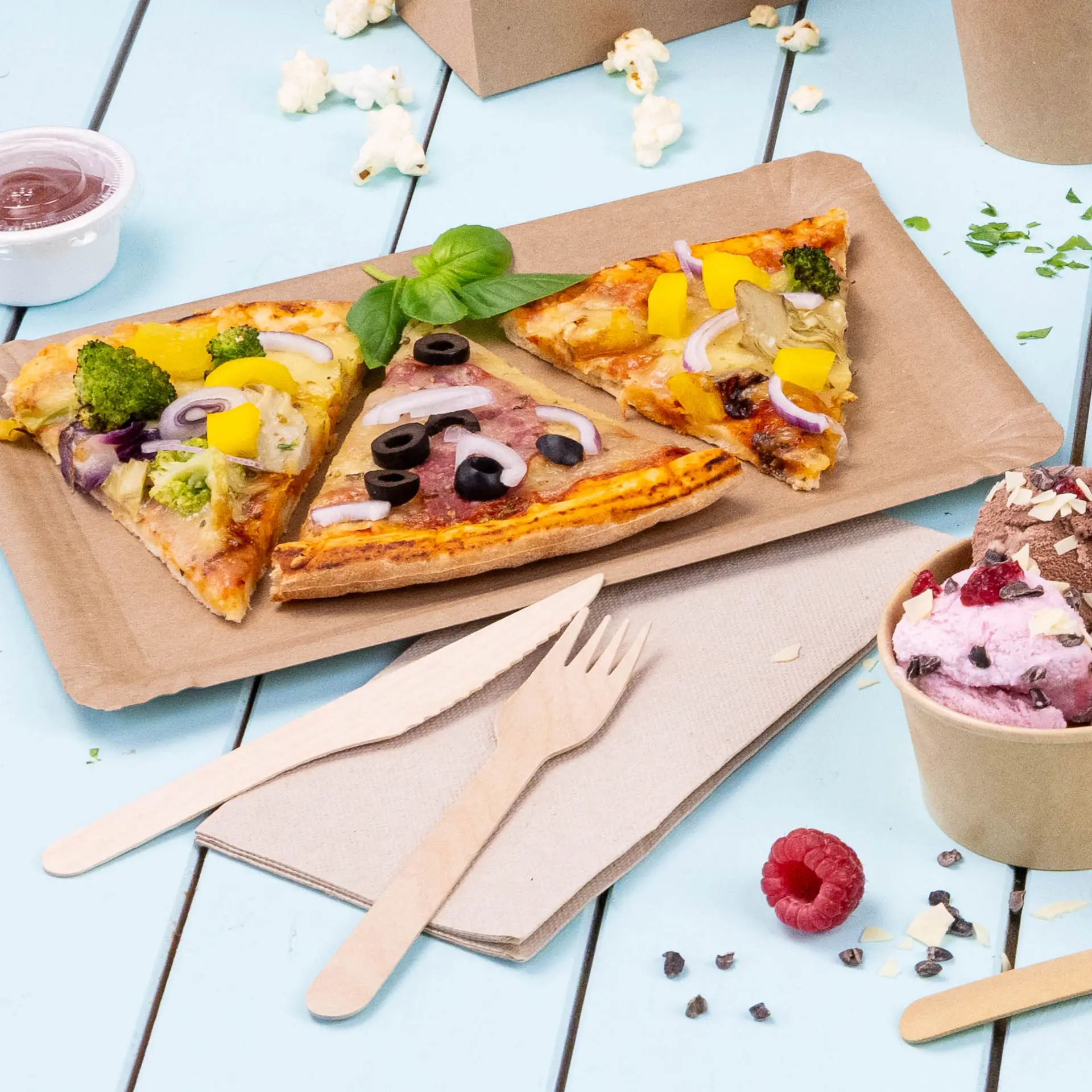 Afbeelding van drie pizzapunten met verschillende toppings op een bruine papieren bord, vergezeld van houten bestek en een beker ijs.