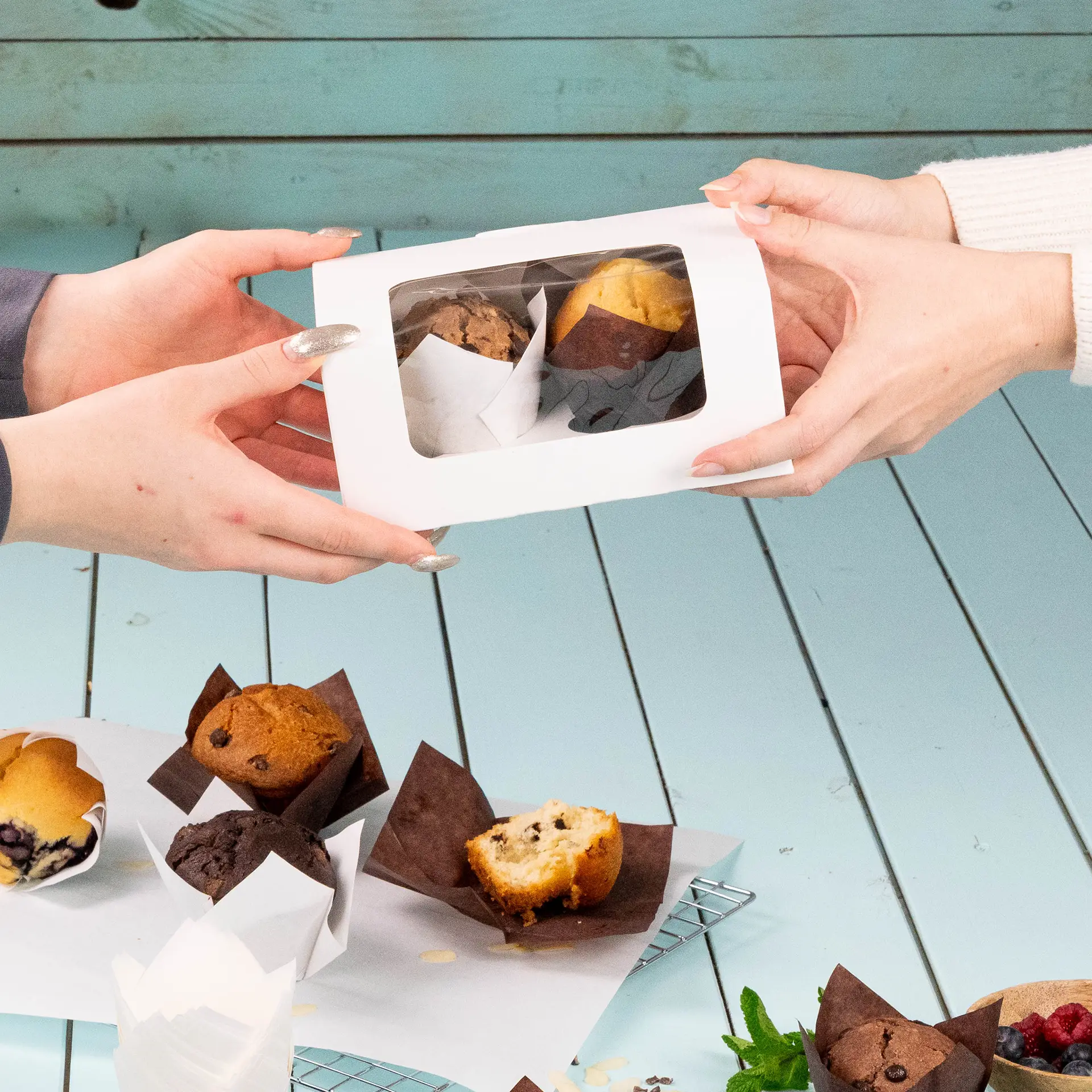 Handen wisselen een doorzichtige doos met twee muffins, met meerdere muffins in bruine cups op een tafel.