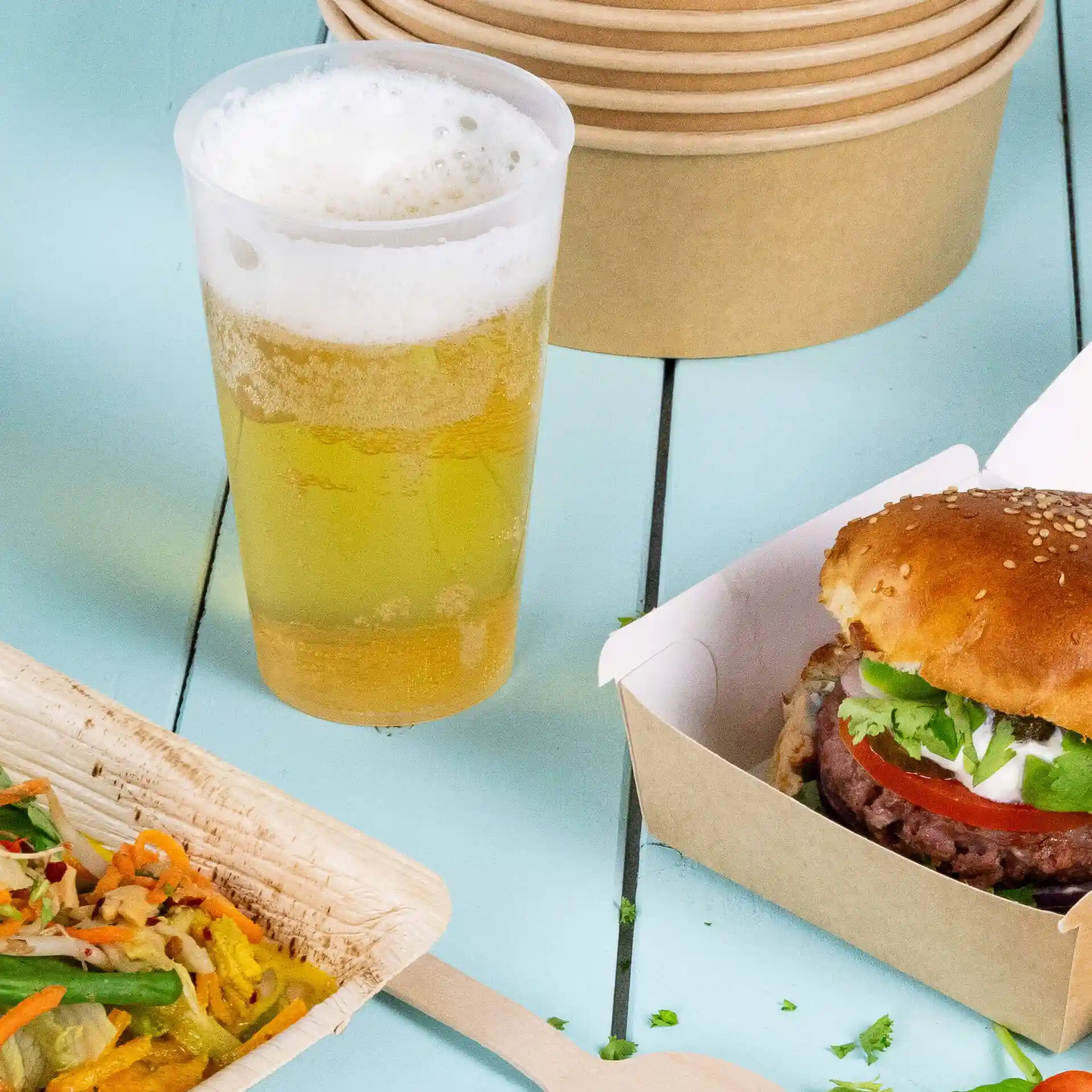 Afbeelding van een transparante herbruikbare beker met bier, een sesamburger in een doos en een houten bord met salade.