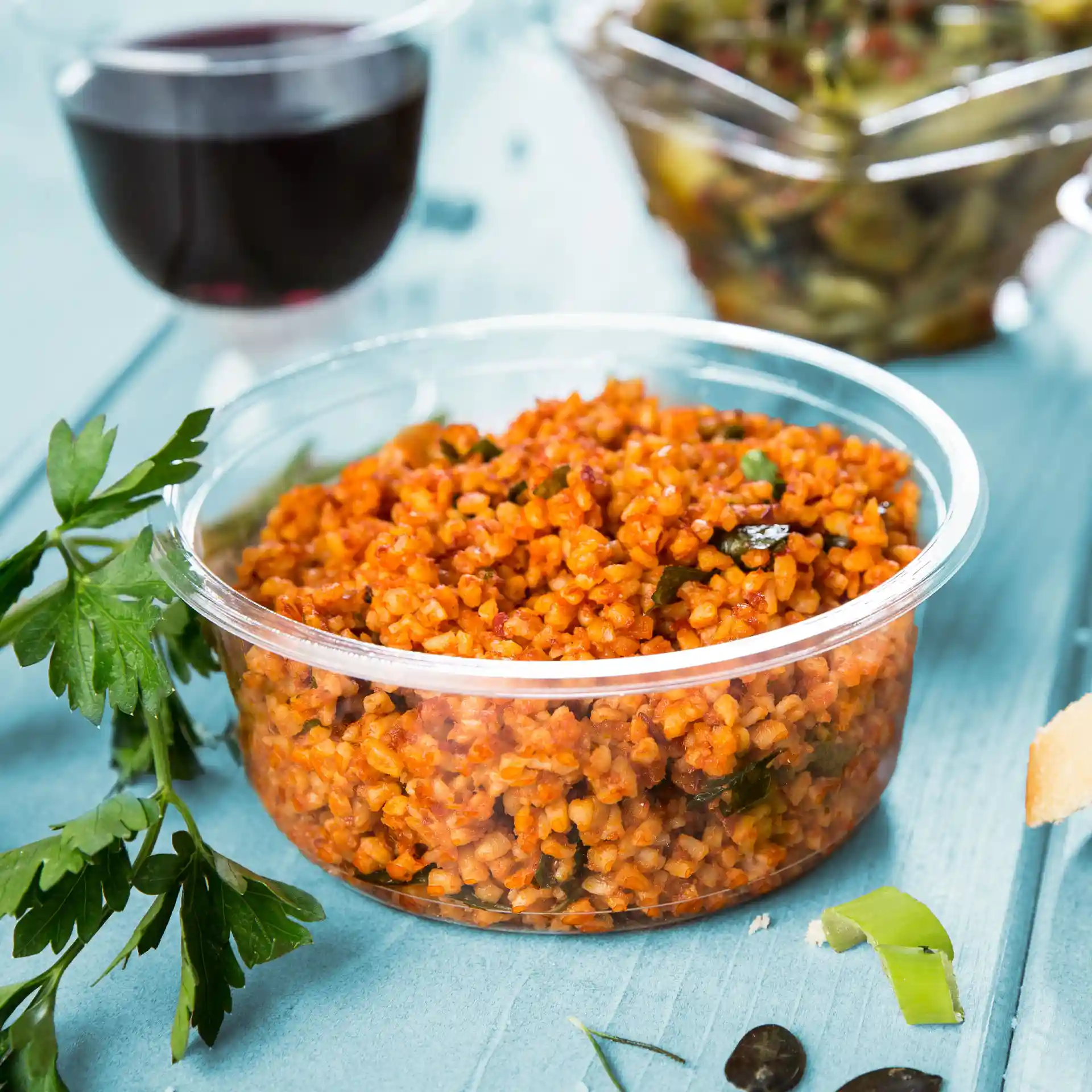 Ronde transparante deli-container met bulgur-gerecht, gegarneerd met kruiden, op een blauwe ondergrond.