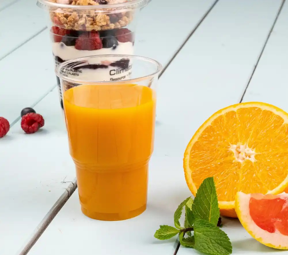 Oranje drank in een transparante beker naast een gehalveerde sinaasappel en muntbladeren, met een dessertbeker op de achtergrond.