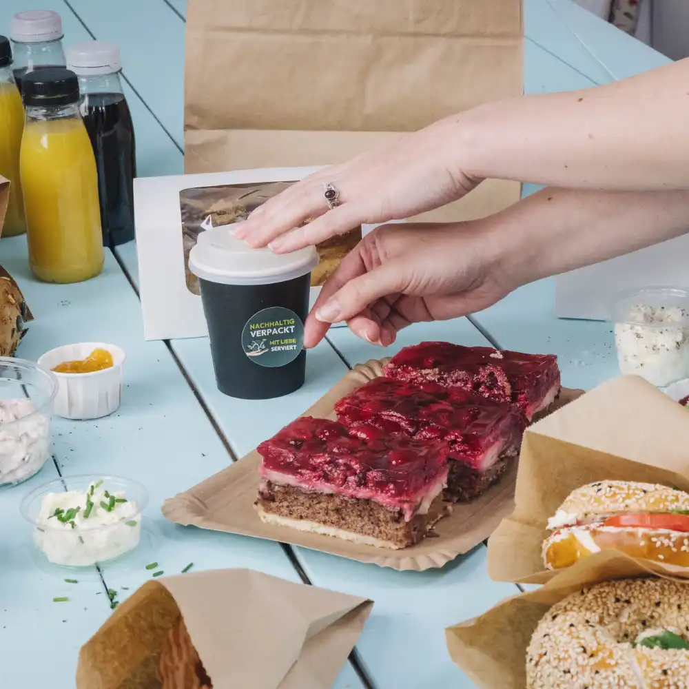 Een hand reikt naar een kleine zwarte drinkbeker op een tafel met verschillende etenswaren; laagjescake, sauzen en drankjes in de buurt.