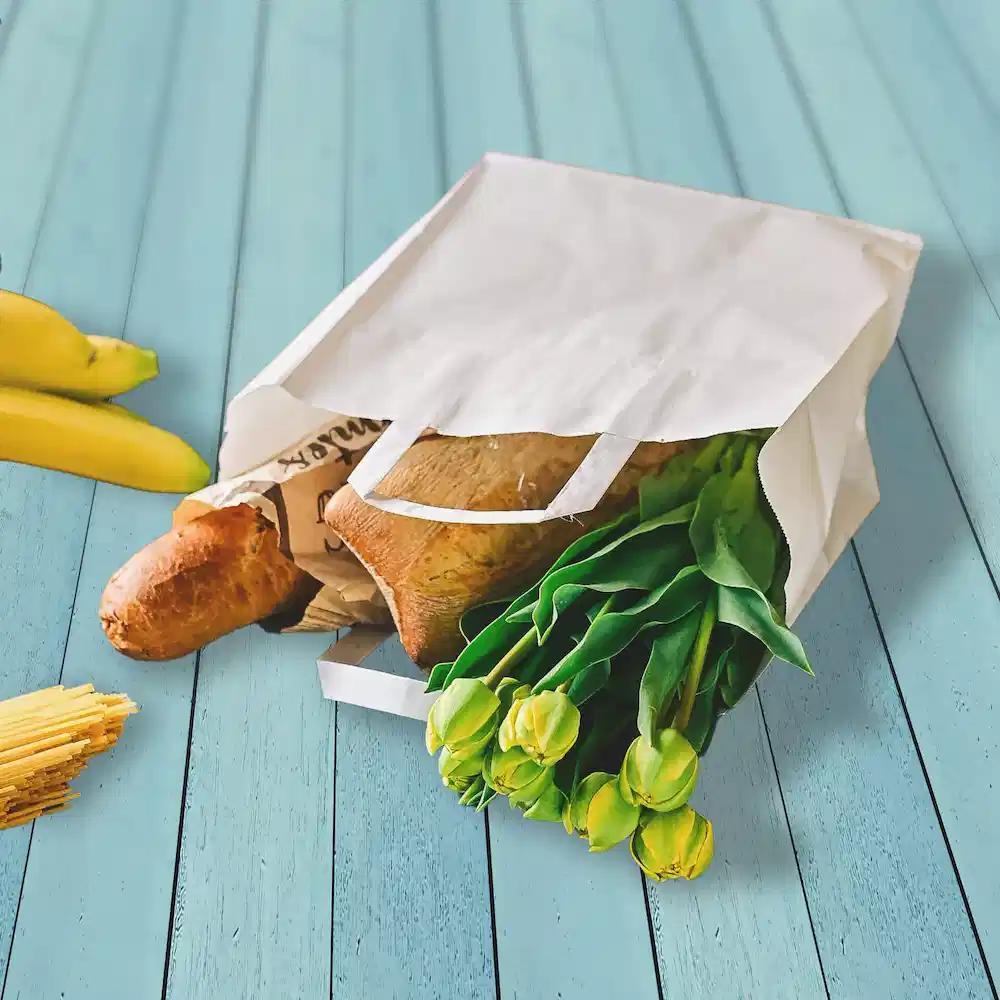 Witte papieren tas gevuld met boodschappen, waaronder brood, tulpen en bananen op een blauwe houten ondergrond.