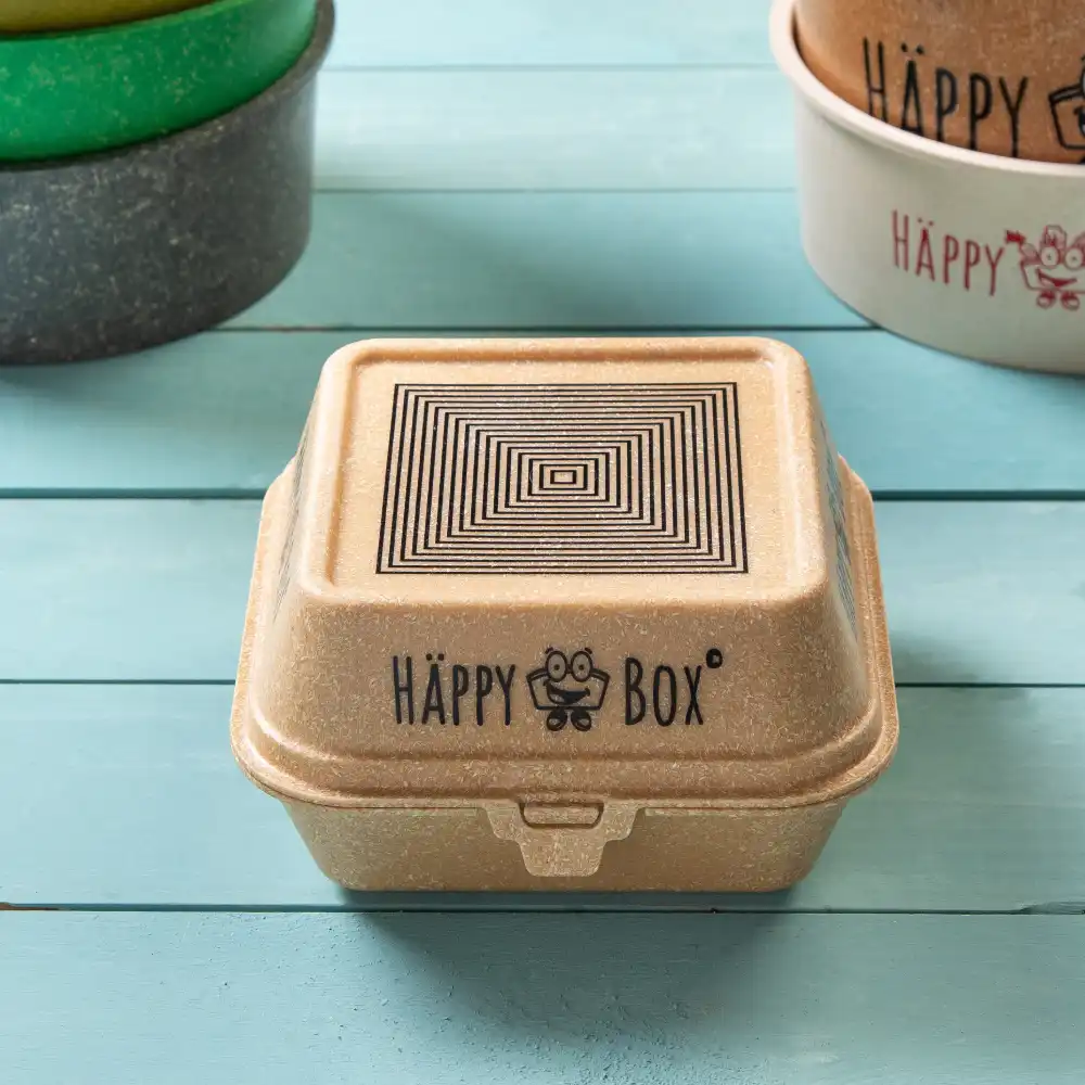 Vierkante veganistische voedselcontainer met een zwart geometrisch ontwerp op het deksel en het "HÄPPY BOX"-logo op een houten oppervlak.