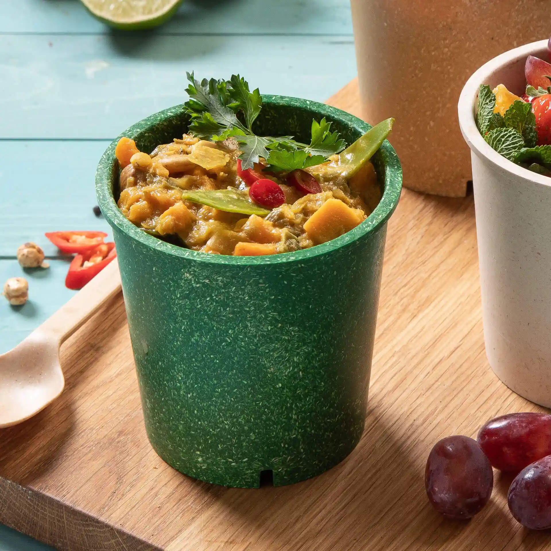 Groene herbruikbare beker gevuld met voedsel, getopt met koriander en rode chili, geplaatst op een houten oppervlak met andere items.
