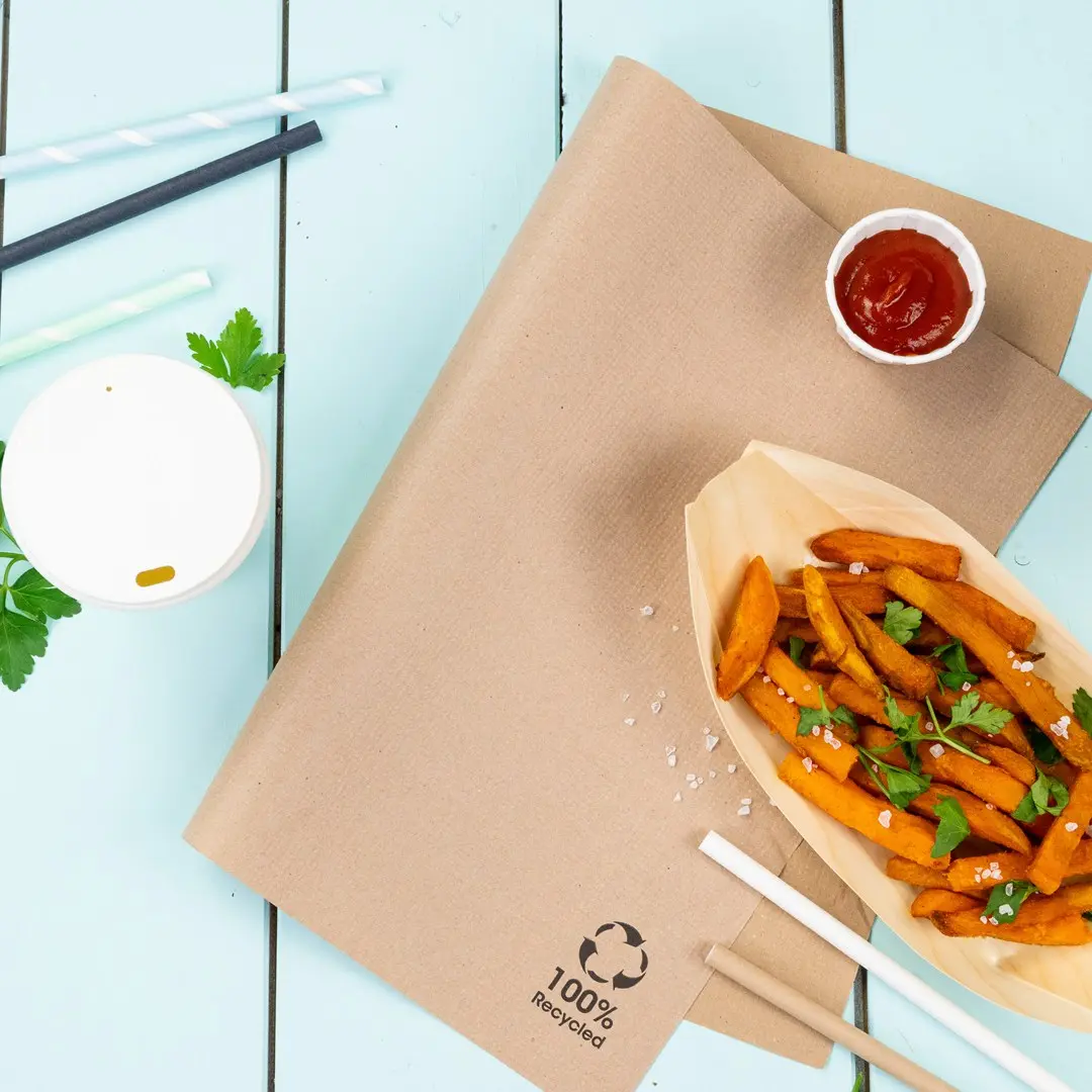 Bruin kraftpapier met zoete aardappelfrites, ketchup, een witte beker en rietjes op een blauwe houten ondergrond.