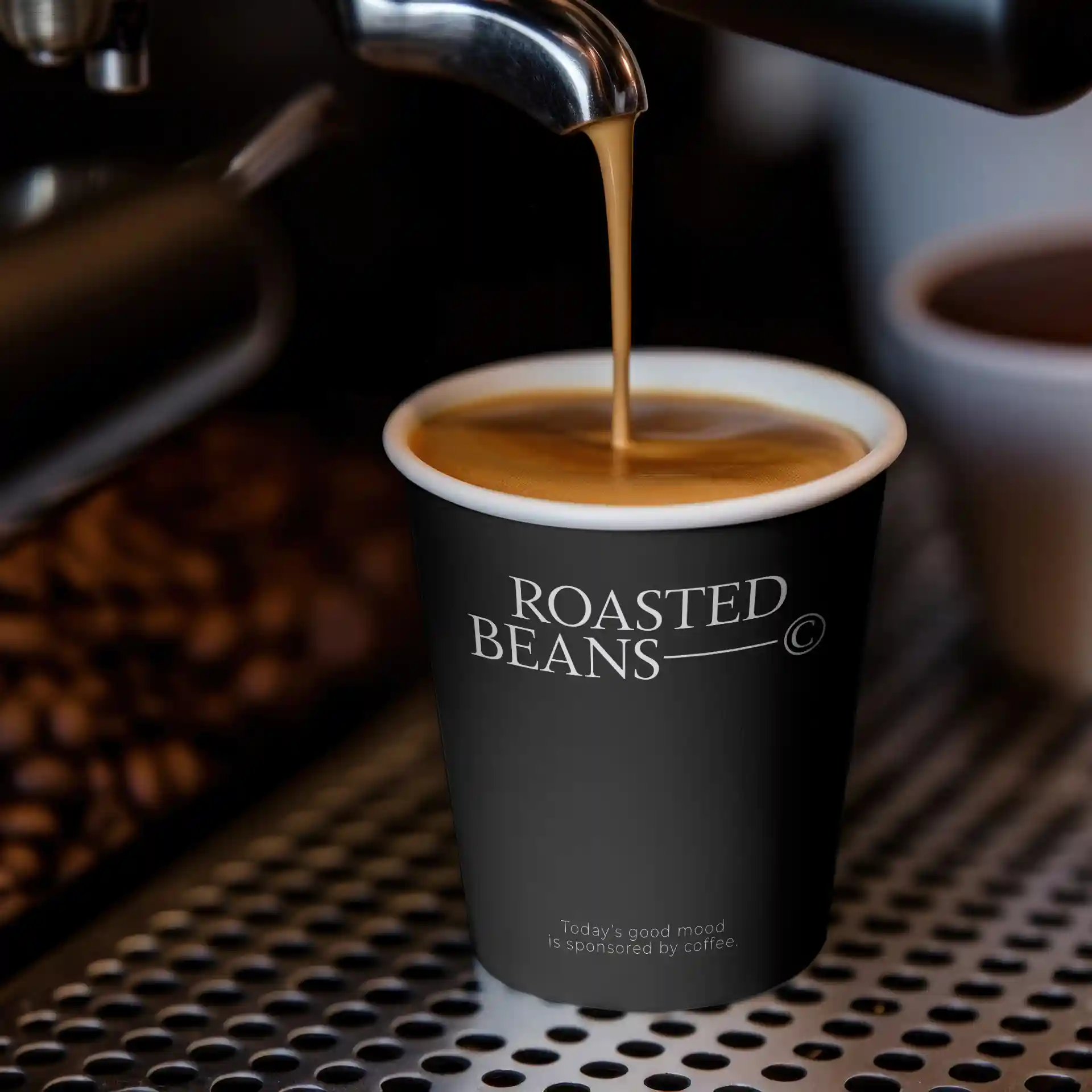 Zwarte papieren beker met het "ROASTED BEANS"-logo, gevuld met koffie, op een metalen oppervlak met koffiebonen op de achtergrond.