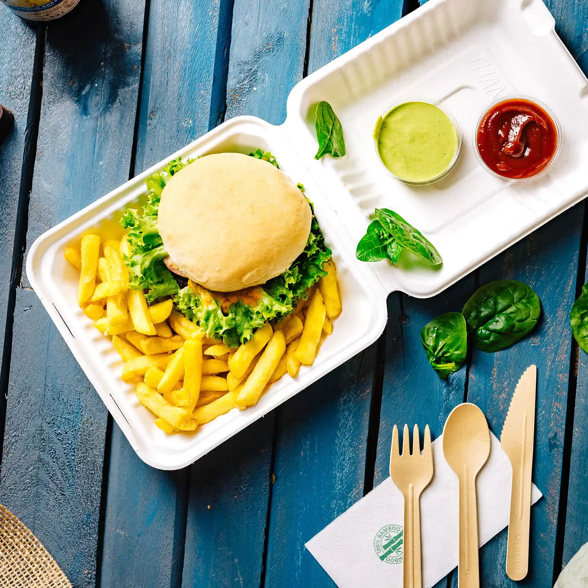 Witte rechthoekige container met scharnierdeksel, met een burger, frietjes, groene saus, ketchup en houten bestek op een blauwe ondergrond.