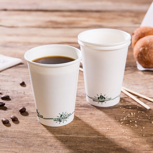 Twee disposable witte papieren bekers op een houten oppervlak; één gevuld met koffie; beide gelabeld met "recyclebaar" en "composteermateriaal".