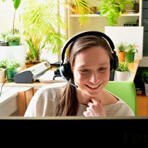 Vrouw met headphones die glimlacht terwijl ze aan een bureau met planten en een computer zit in een lichte kamer.