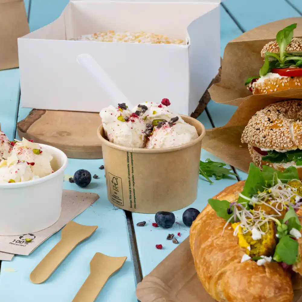 IJs in een beker met toppings, twee sandwiches op papier, een croissant met vulling, en bosbessen op een blauwe houten oppervlak.
