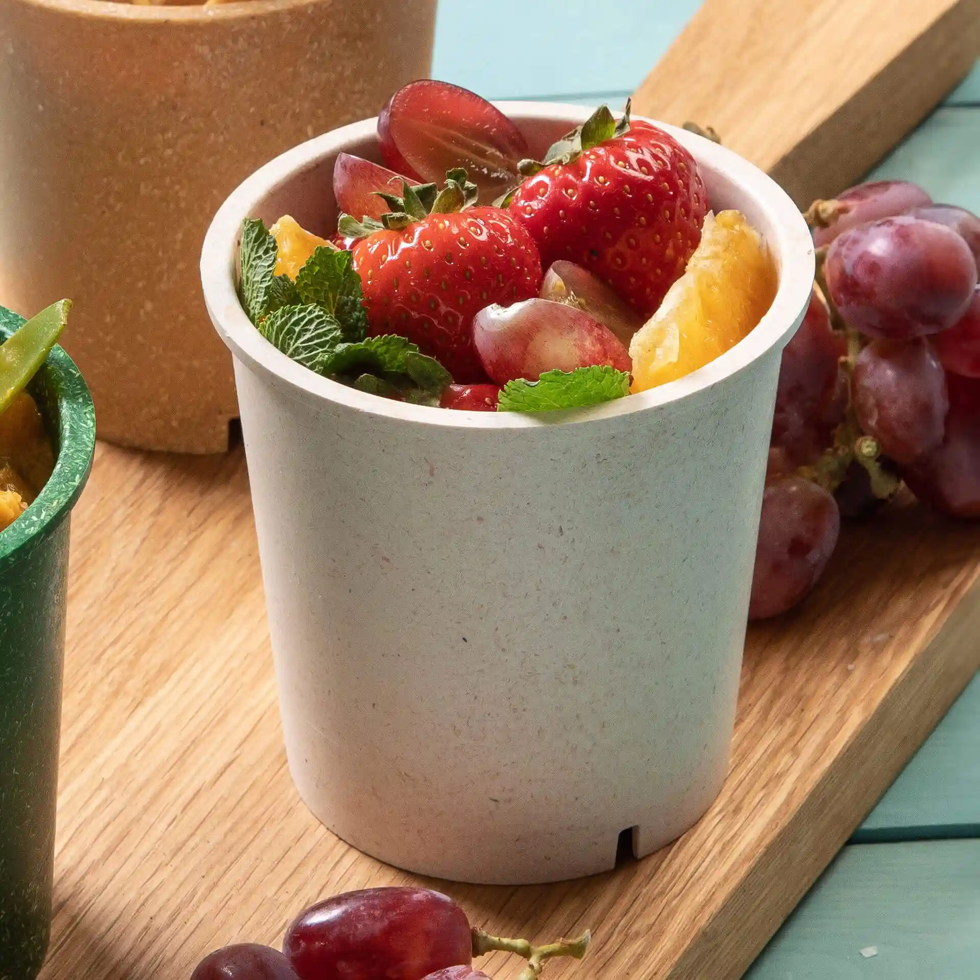 Ronde crème-witte herbruikbare beker gevuld met aardbeien, druiven en muntblaadjes op een houten oppervlak.