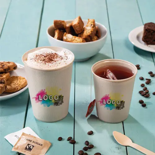 Twee bekers met logo-ontwerp, de ene met slagroom en de andere met ijsthee, omringd door koekjes, taart en suikerzakjes.