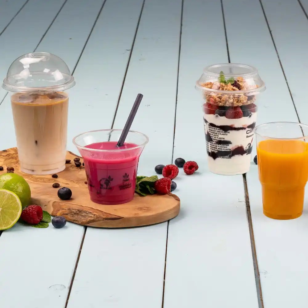 Vier drankjes op een houten plank: iced coffee in een beker met een deksel, roze drankje met een rietje, gelaagd dessertbekertje en sinaasappelsap.