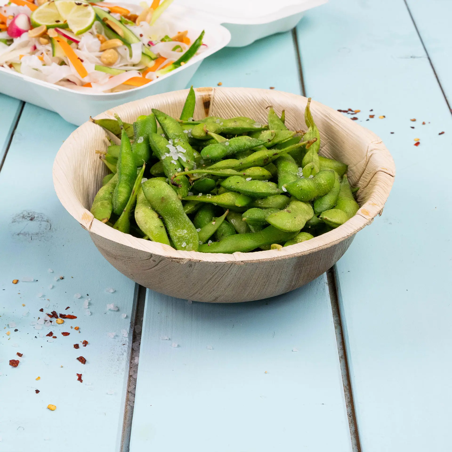 Ronde palmbladkom gevuld met groene edamame, bestrooid met zout, op een blauwe houten ondergrond.