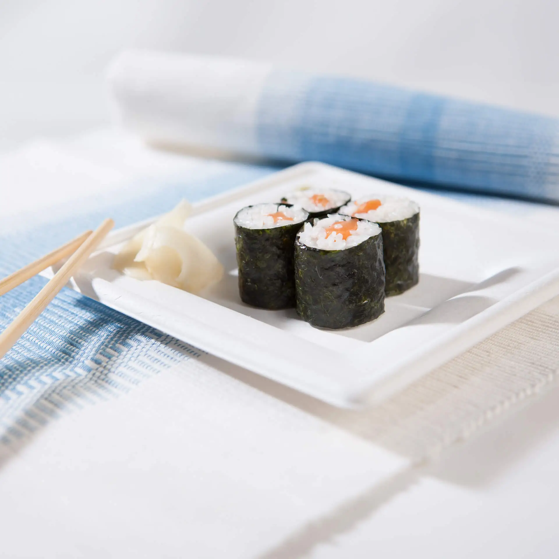 Vierkante suikerrietplaat met sushirollen, ingelegde gember en chopsticks op een gestructureerde stoffen achtergrond.