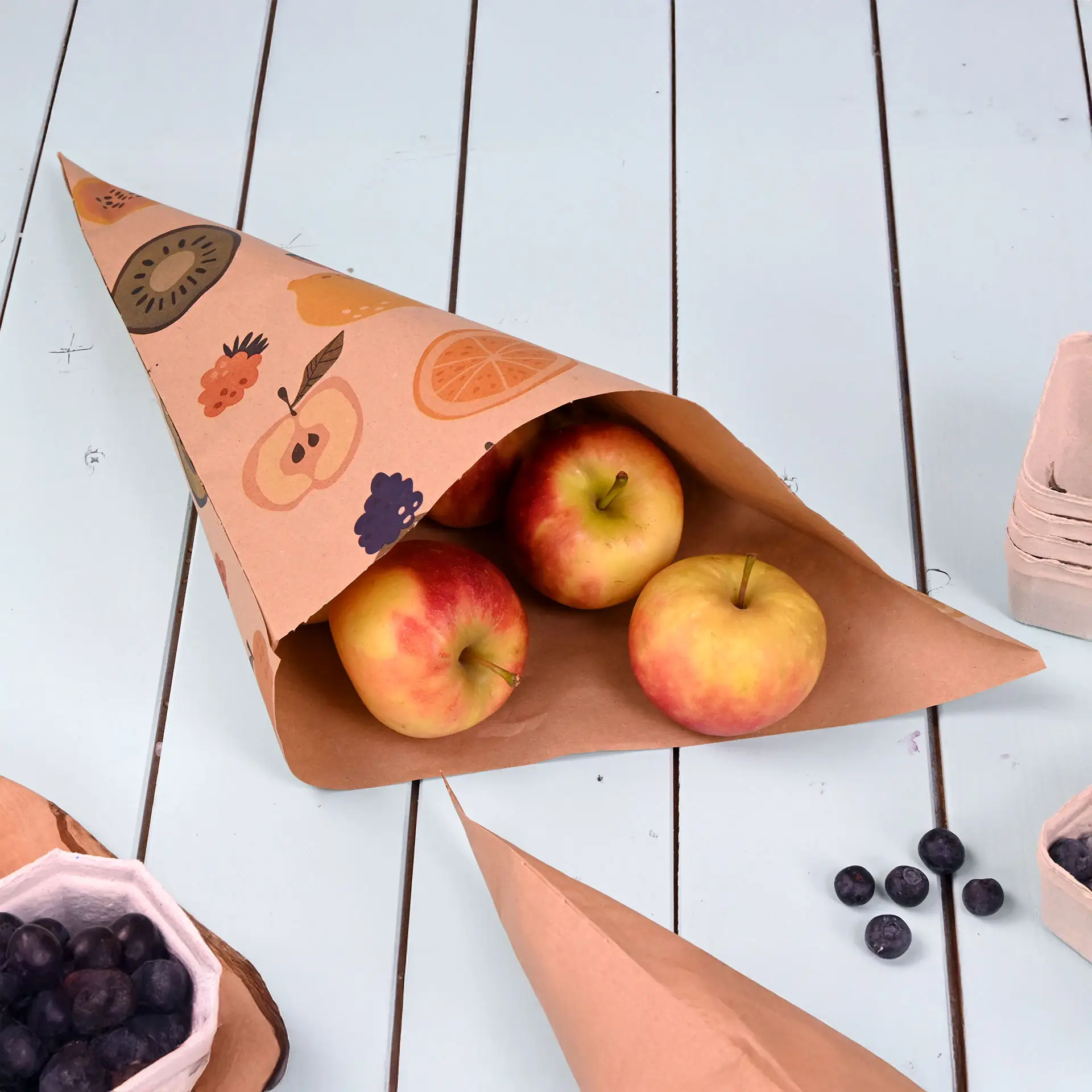 Bruine papieren kegel met fruitdesign, met drie rode appels, op een lichtblauw tafelblad.