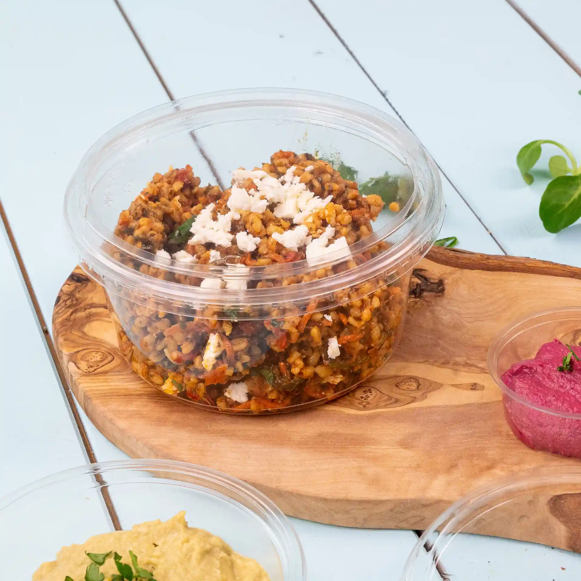 Transparante ronde deli-container met een deksel, gevuld met voedsel en op een houten plank geplaatst.