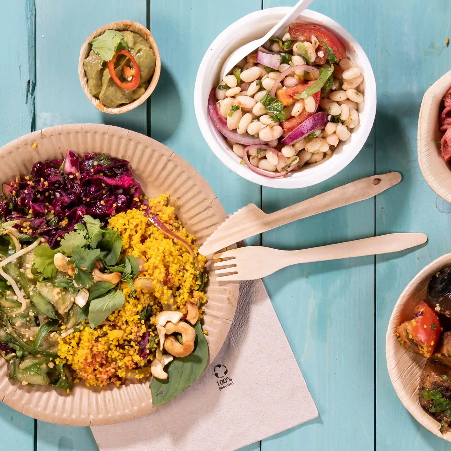 Afbeelding van een bord met kleurrijk voedsel, waaronder couscous, salade en een klein kommetje bonen, vergezeld van houten vorken op een blauwe ondergrond.