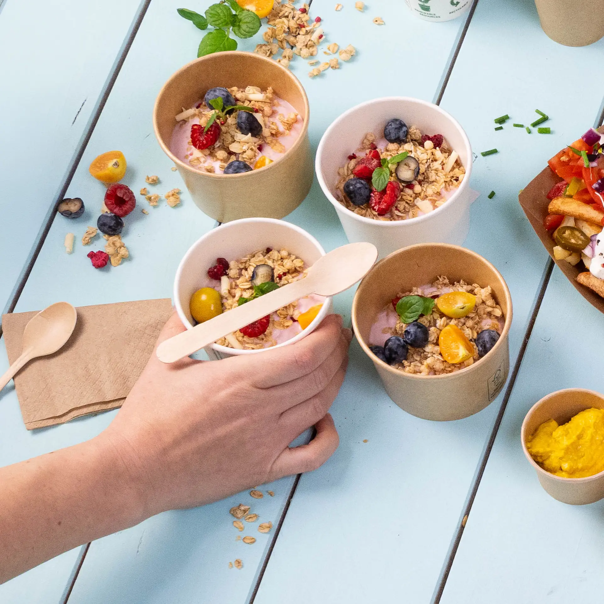 Afbeelding van verschillende kleine bekers met yoghurt, getopt met granola en fruit, een hand die een beker vasthoudt, en houten lepels in de buurt.