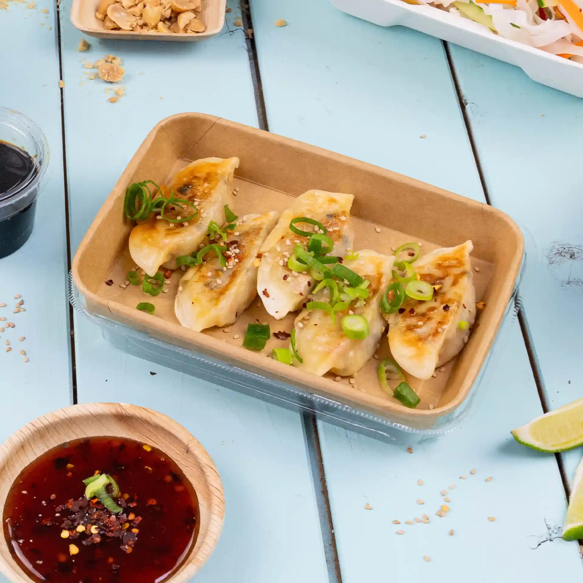Container met vijf dumplings, getopt met lente-ui en sesam, vergezeld van een kom dipsaus.
