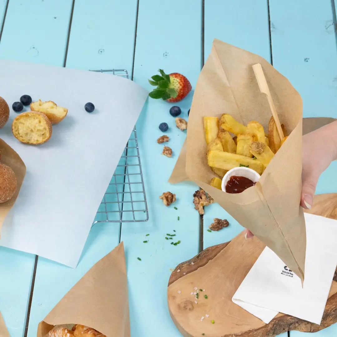 Bruine papieren kegel gevuld met frietjes en een klein bekertje ketchup, omringd door gebak, bessen en een houten plank.