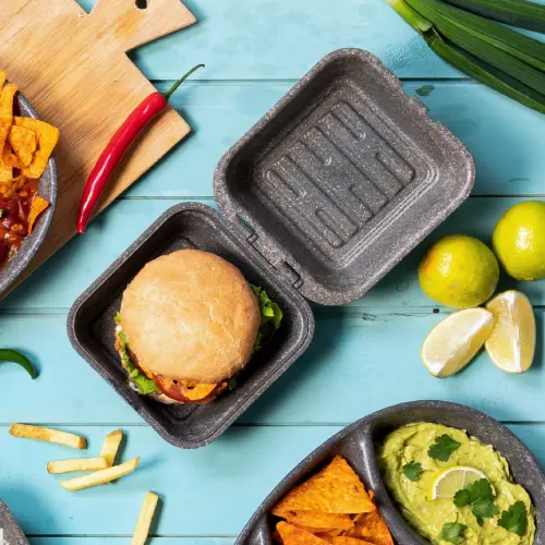 Burger in een herbruikbare kraftdoos met sla en sauzen, omringd door chips, frietjes, guacamole en limoenpartjes.