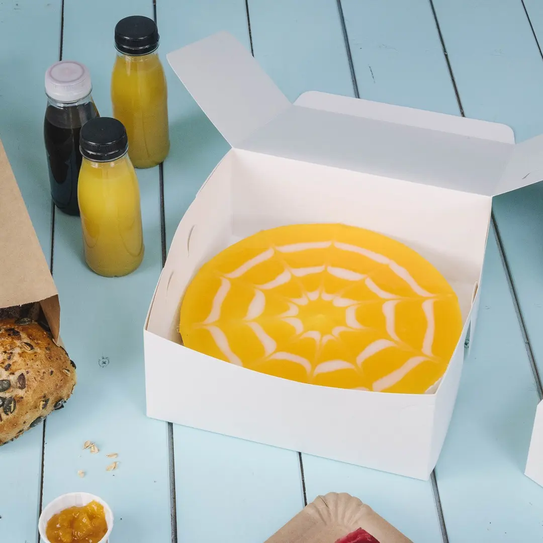 Witte cakebox met een gele cake en een wit spinnenwebontwerp, omringd door dranken en gebakken goederen.