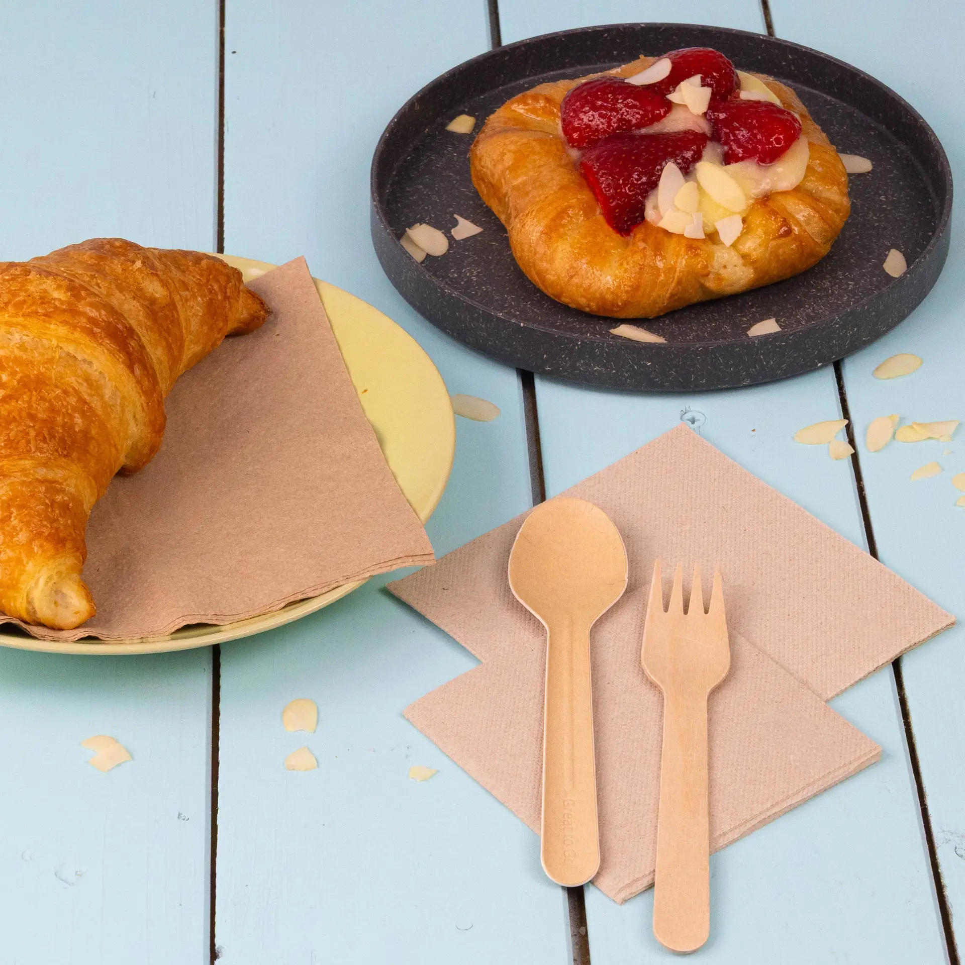 Afbeelding van een croissant op een gele plaat, een fruitgebak op een zwarte plaat en gerecyclede servetten met een lepel en vork.