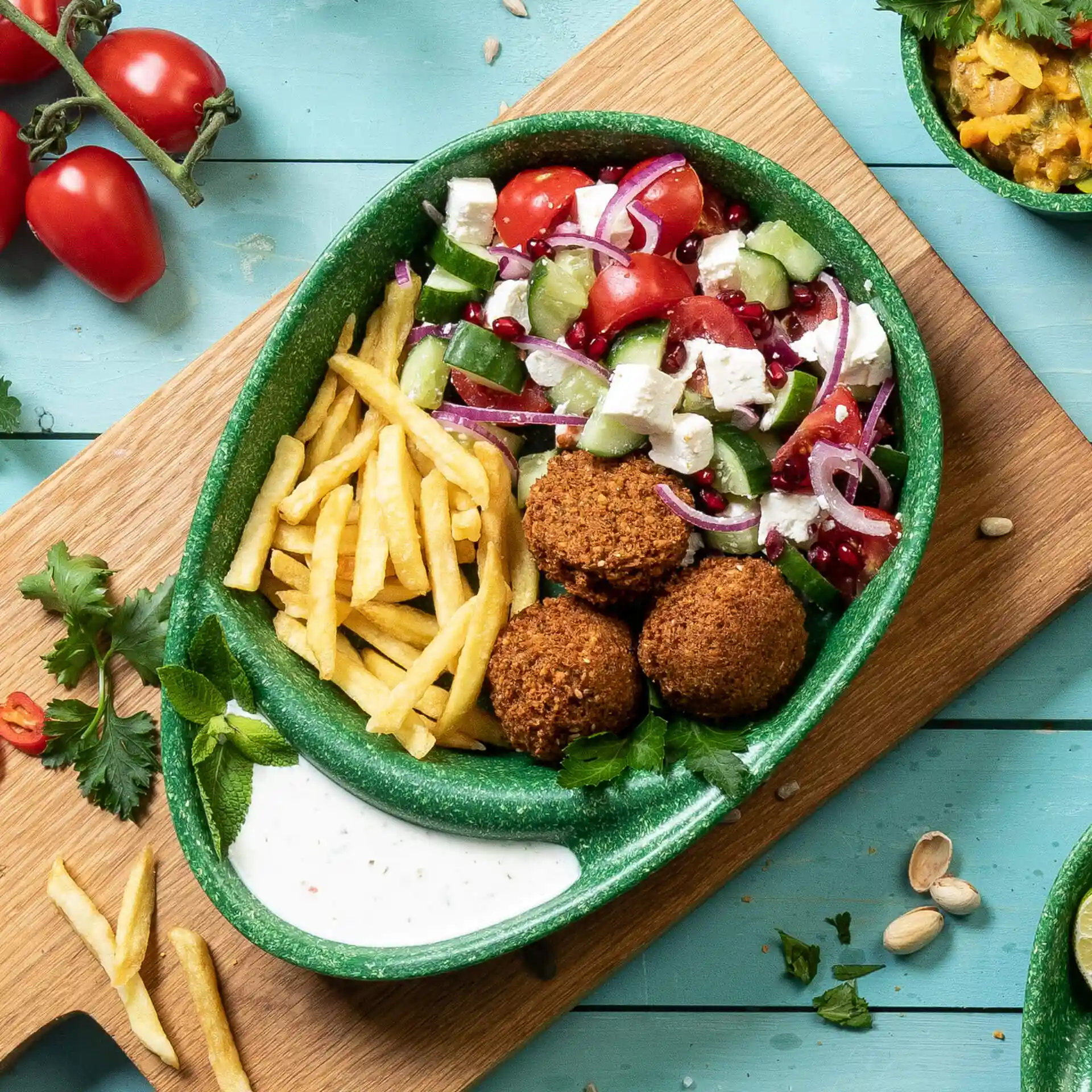Groen verdeeld bord met falafelballen, frietjes, salade en saus, geplaatst op een houten oppervlak met tomaten en kruiden in de buurt.