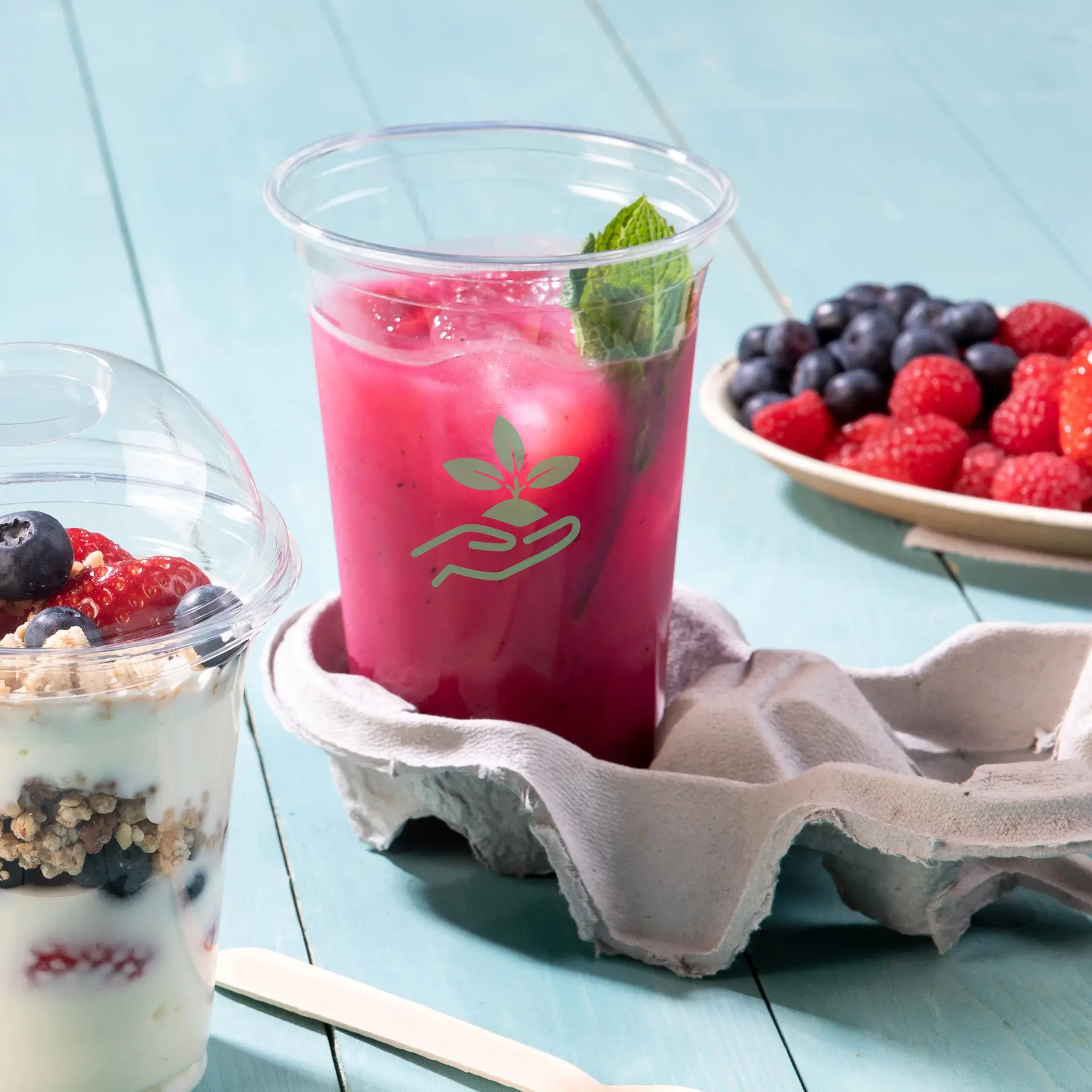 Een doorzichtig rPET-bekertje gevuld met een roze drankje en een muntblad; een eierdoos houdt het bekertje vast; een yoghurtparfait en bessen zijn in de buurt.