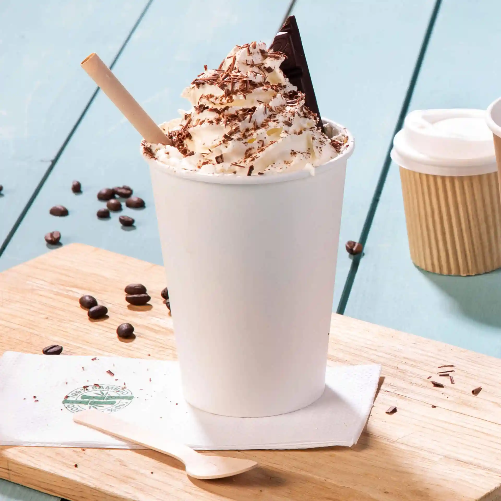 Witte beker gevuld met slagroom, chocoladeschilfers en een stuk chocolade, met een houten lepel ernaast.