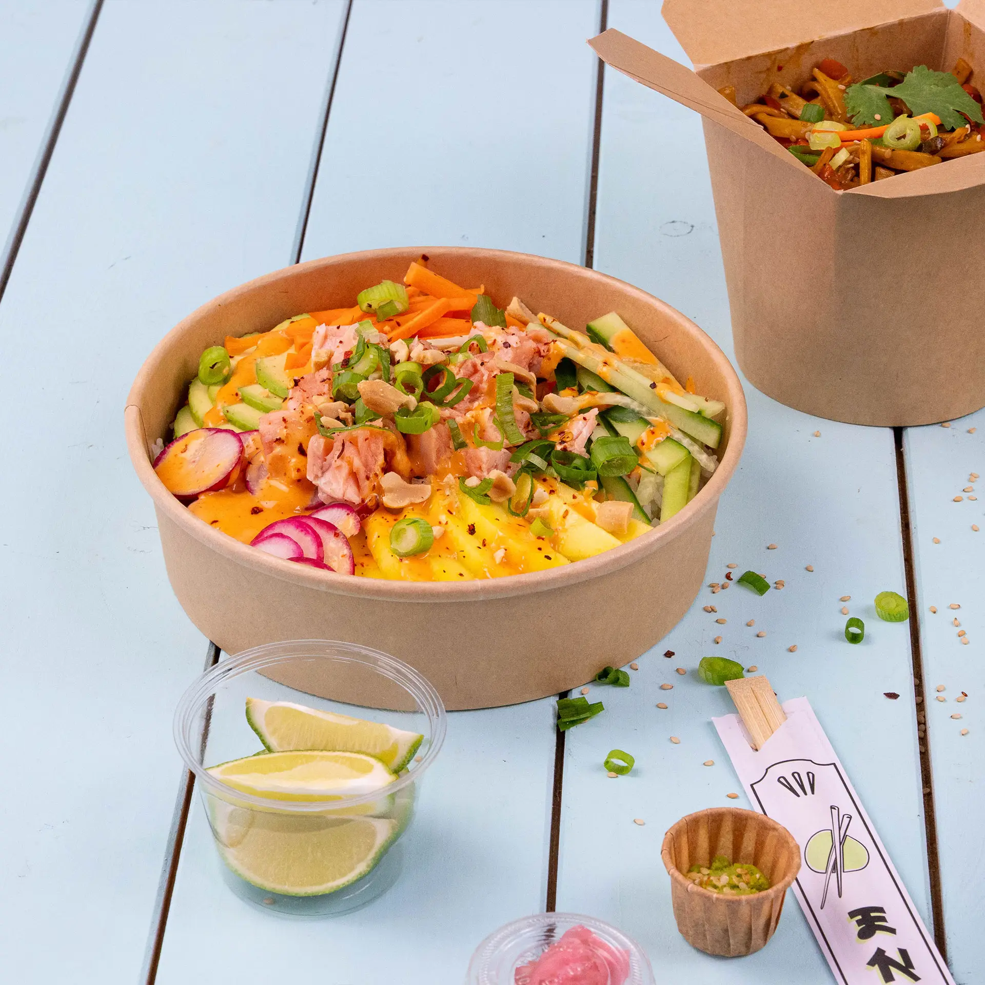 Bruine papieren saladekom gevuld met gemengde groenten en toppings, met limoenschijfjes en een tweede container op de achtergrond.