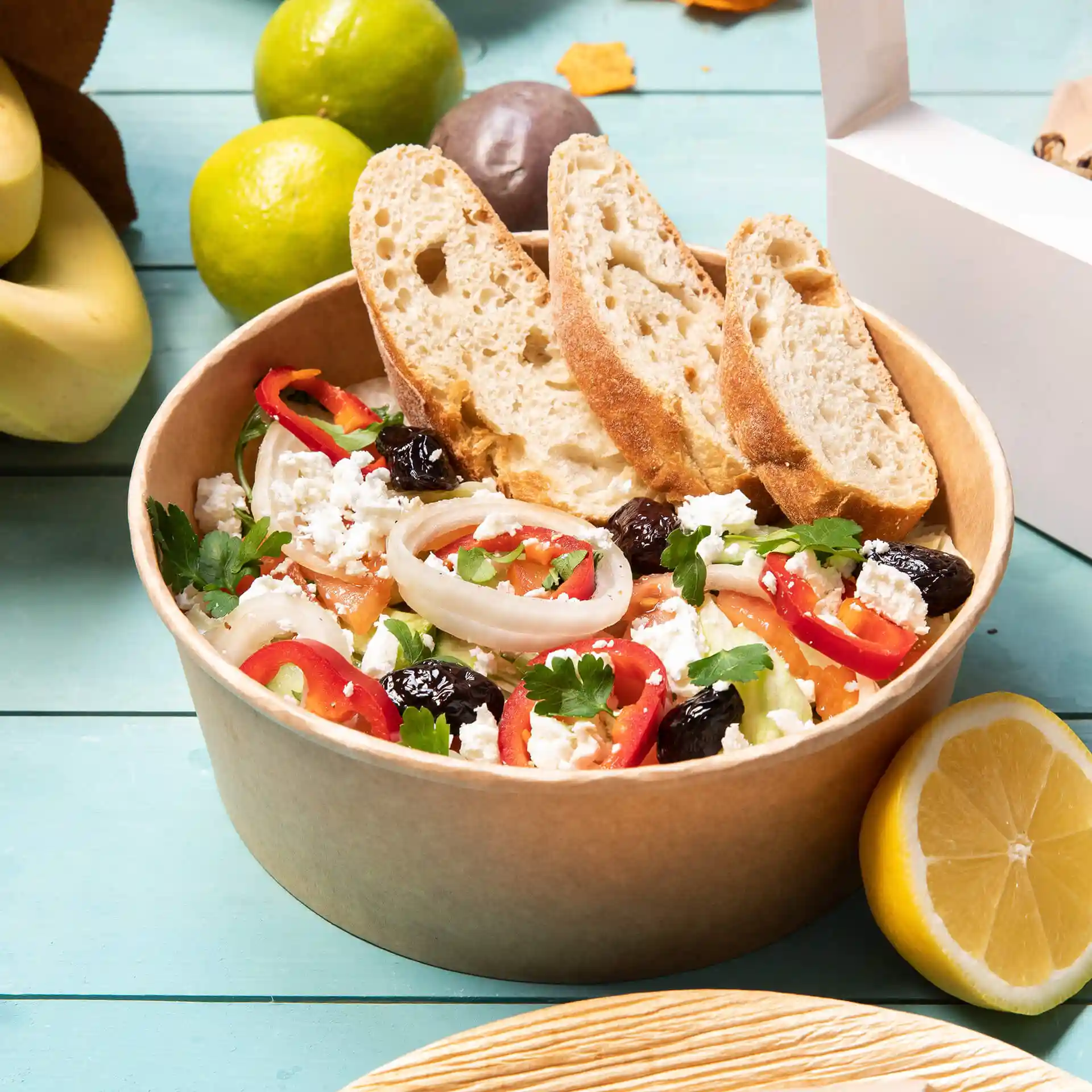 Kom gevuld met salade, getopt met gesneden groenten en feta, vergezeld van twee sneetjes brood, op een blauwe ondergrond.