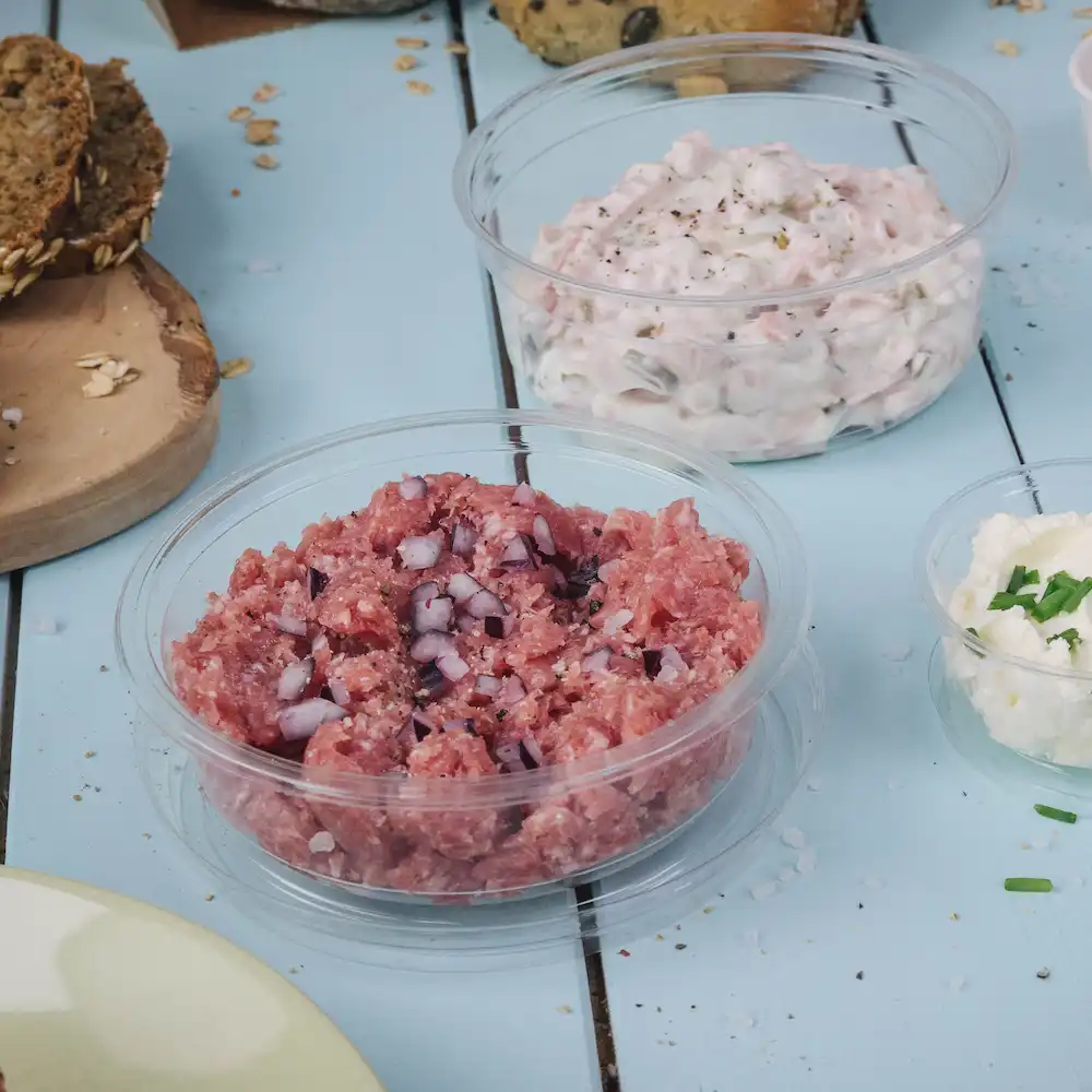 In blokjes gesneden rauw vlees met ui in een doorzichtige container; daarnaast een romige dip in een andere container en een klein schaaltje kaas.