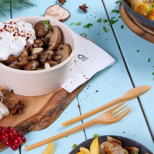 Kom met gebakken paddenstoelen, getopt met yoghurt en specerijen, vergezeld van houten bestek op een blauwe achtergrond.