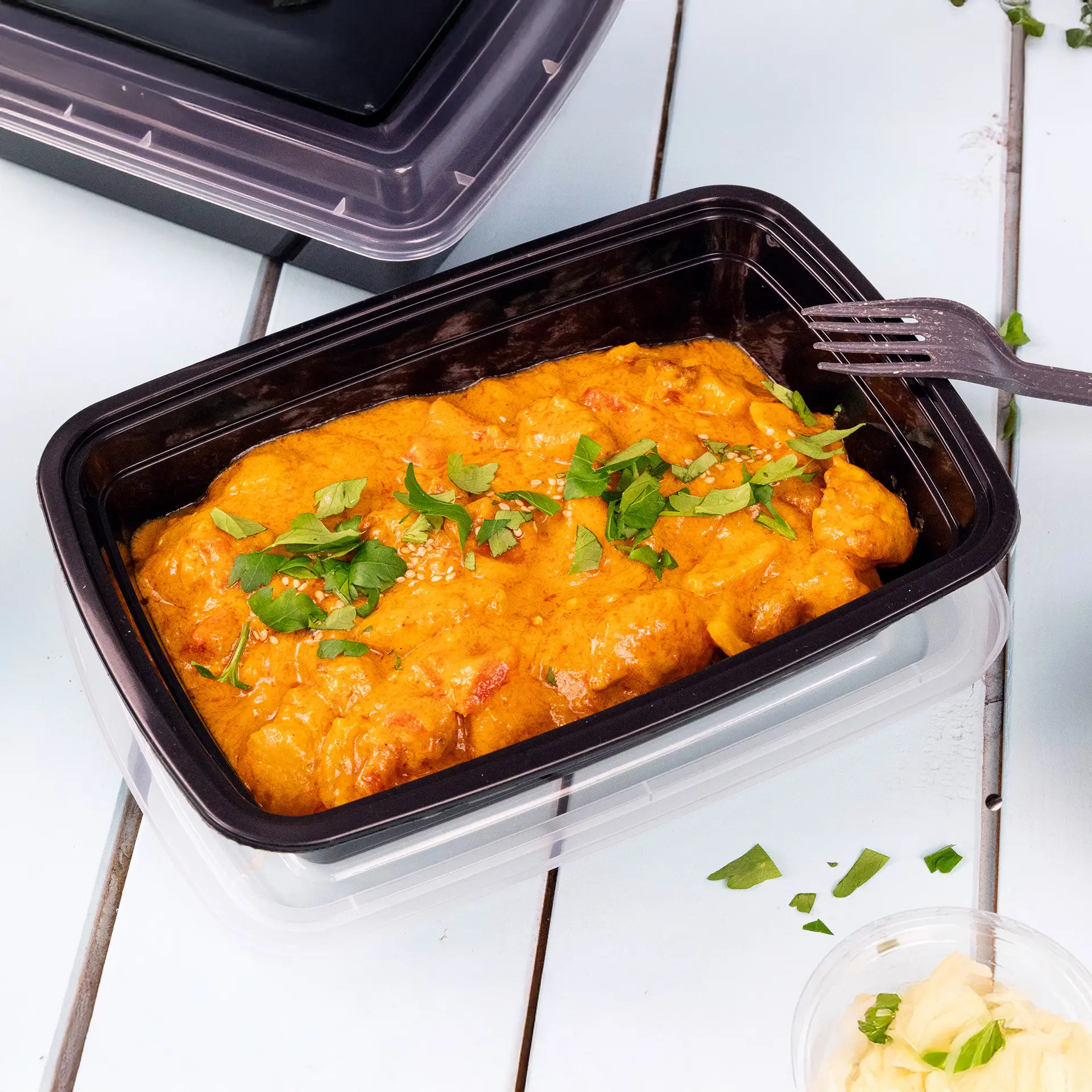 Herbruikbare zwarte polypropyleen voedselcontainer met currygerecht, gegarneerd met kruiden; deksel en vork in de buurt.