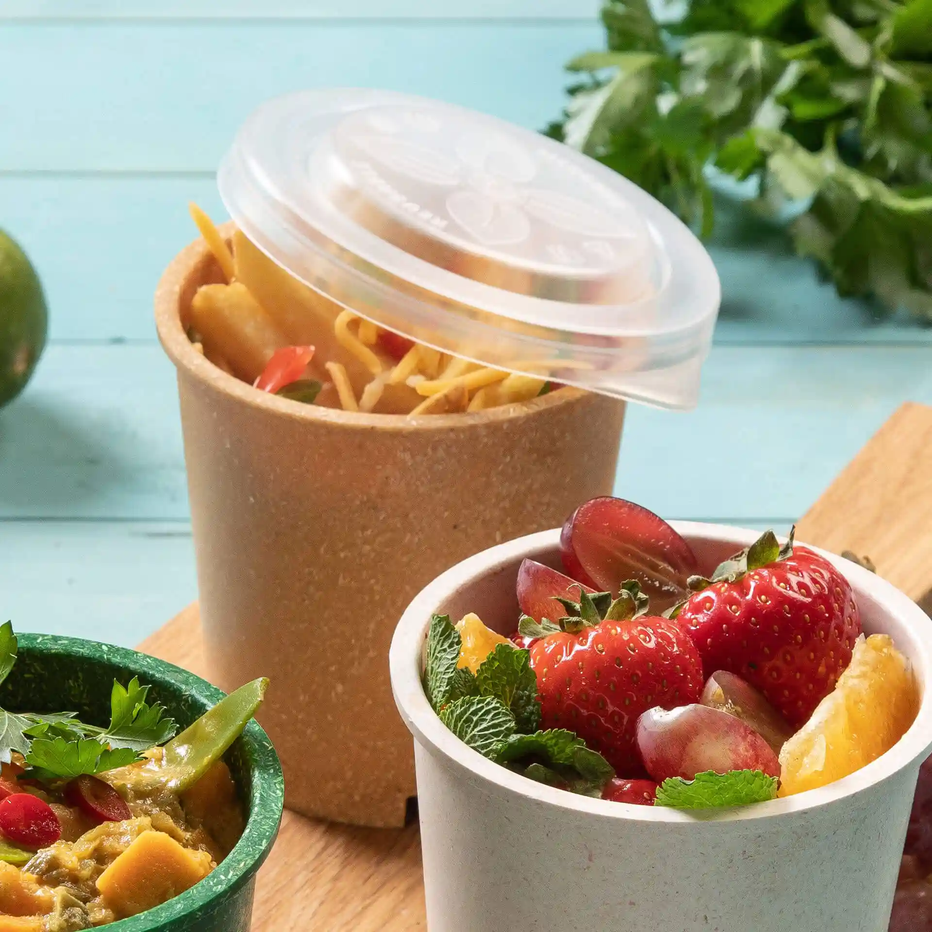 Afbeelding van twee ronde bekers, de ene met een transparant deksel met noedels, de andere met fruit, op een houten oppervlak.
