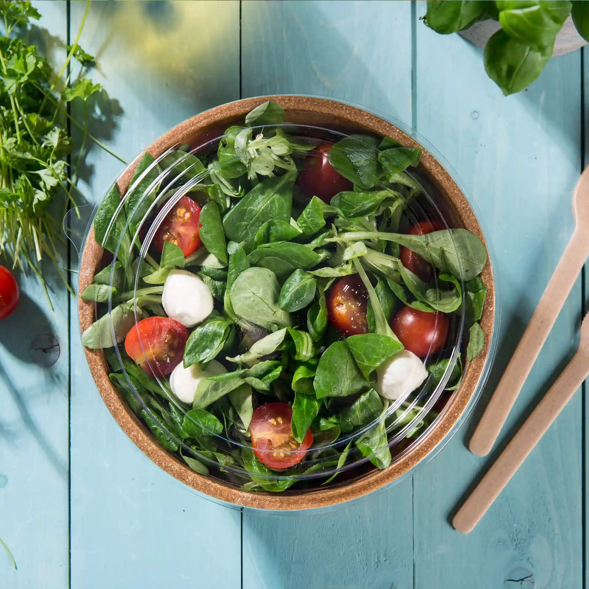 Duidelijk rond deksel van PLA, geplaatst op een kom salade met tomaten en spinazie op een blauwe houten ondergrond.