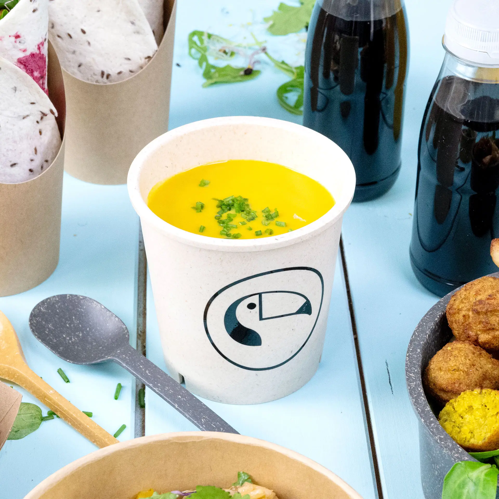Crèmewitte herbruikbare soepbeker gevuld met gele soep en getopt met bieslook, op een blauwe achtergrond met ander eten.