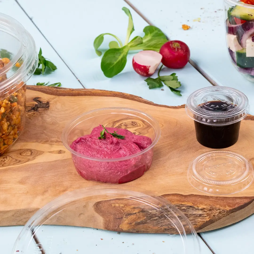 Heldere plastic portie beker met roze dip, omringd door een houten plank, salade-ingrediënten en een andere kleine beker.