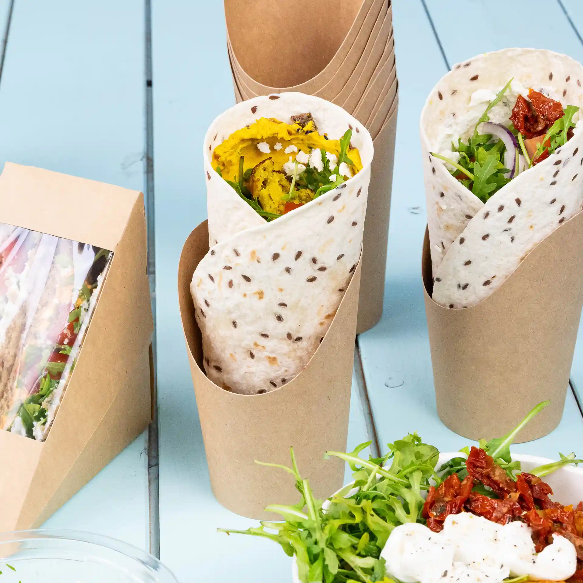 Afbeelding van twee wrapbekers van bruin karton, één gevuld met eten en een bijsalade met groene bladeren en zongedroogde tomaten.