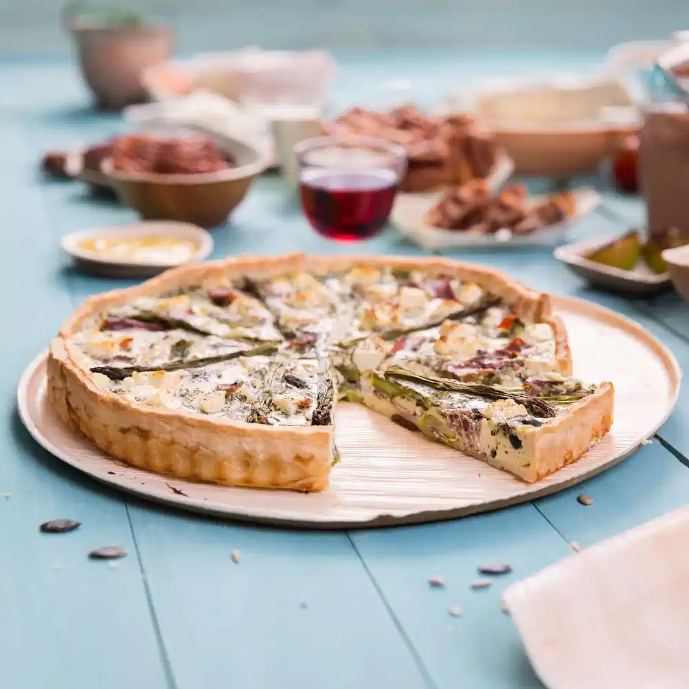 Hartige quiche op een houten bord, met een stuk eruit, omringd door verschillende gerechten en een glas rode drank.
