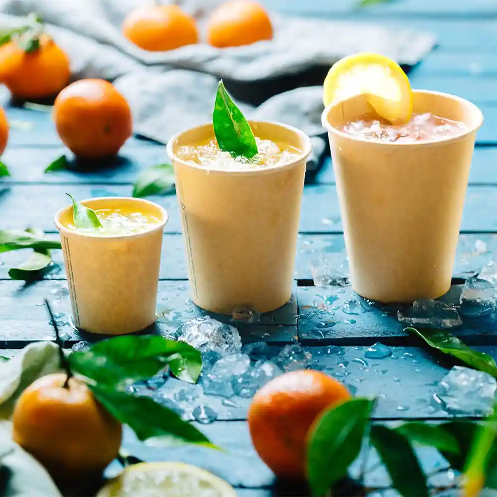 Drie bruine papieren bekers gevuld met drankjes, op een blauwe houten ondergrond met ijs en citrusvruchten eromheen.