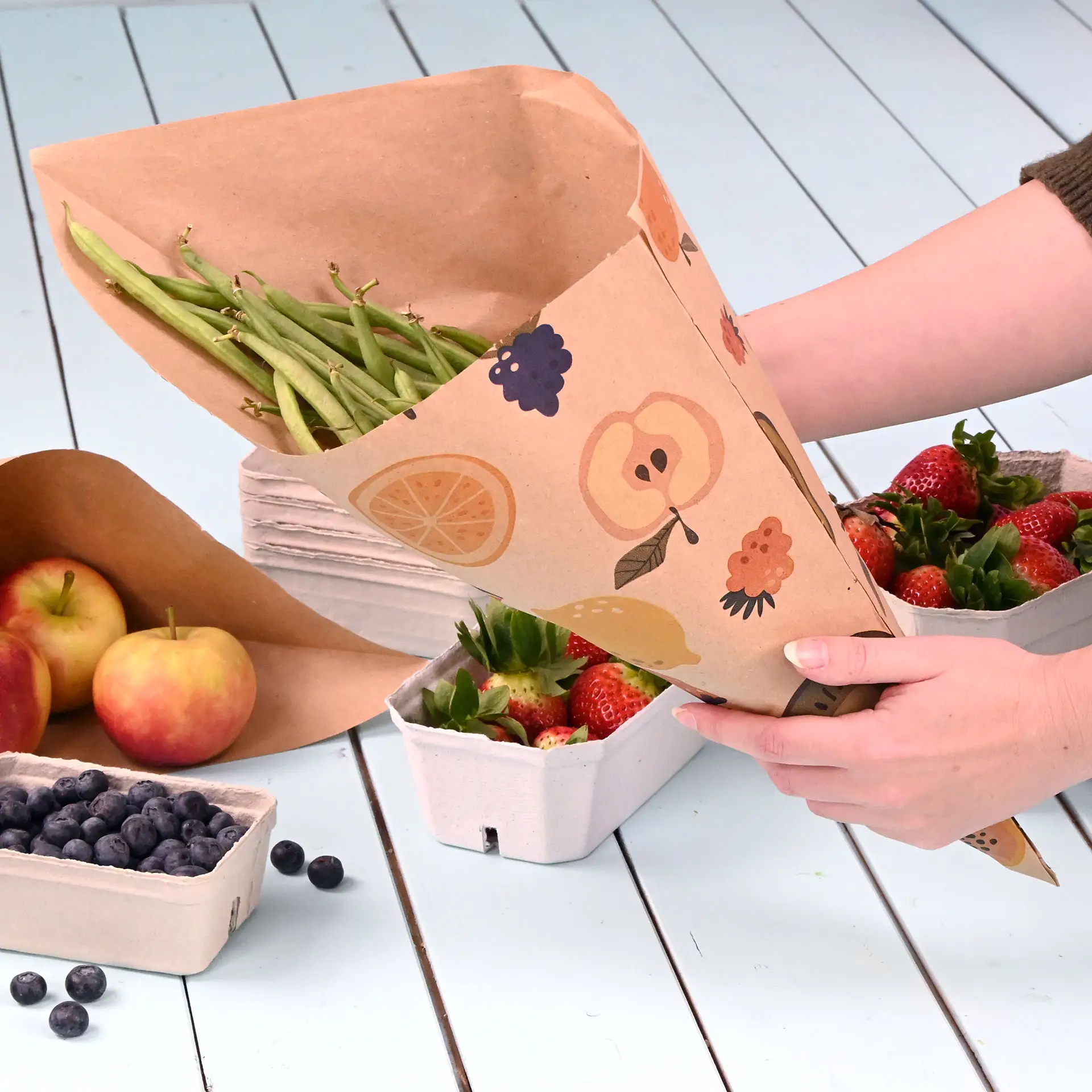 Een hand houdt een bruine papieren cone gevuld met groene bonen, omringd door aardbeien, appels en blauwe bessen op een lichtblauwe ondergrond.