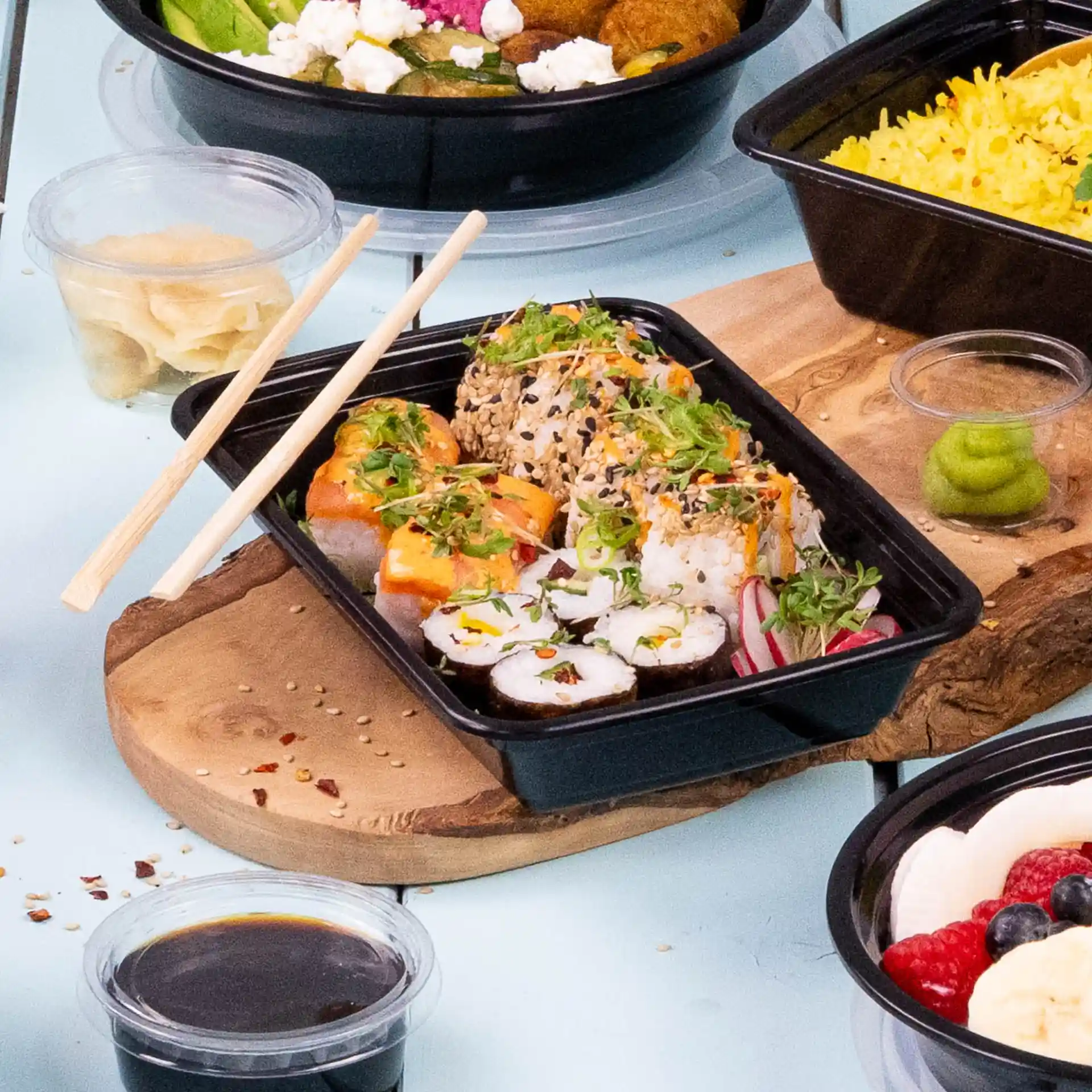 Sushirollen met toppings in een zwarte rechthoekige herbruikbare container, stokjes ernaast en sauzen zoals wasabi en sojasaus.