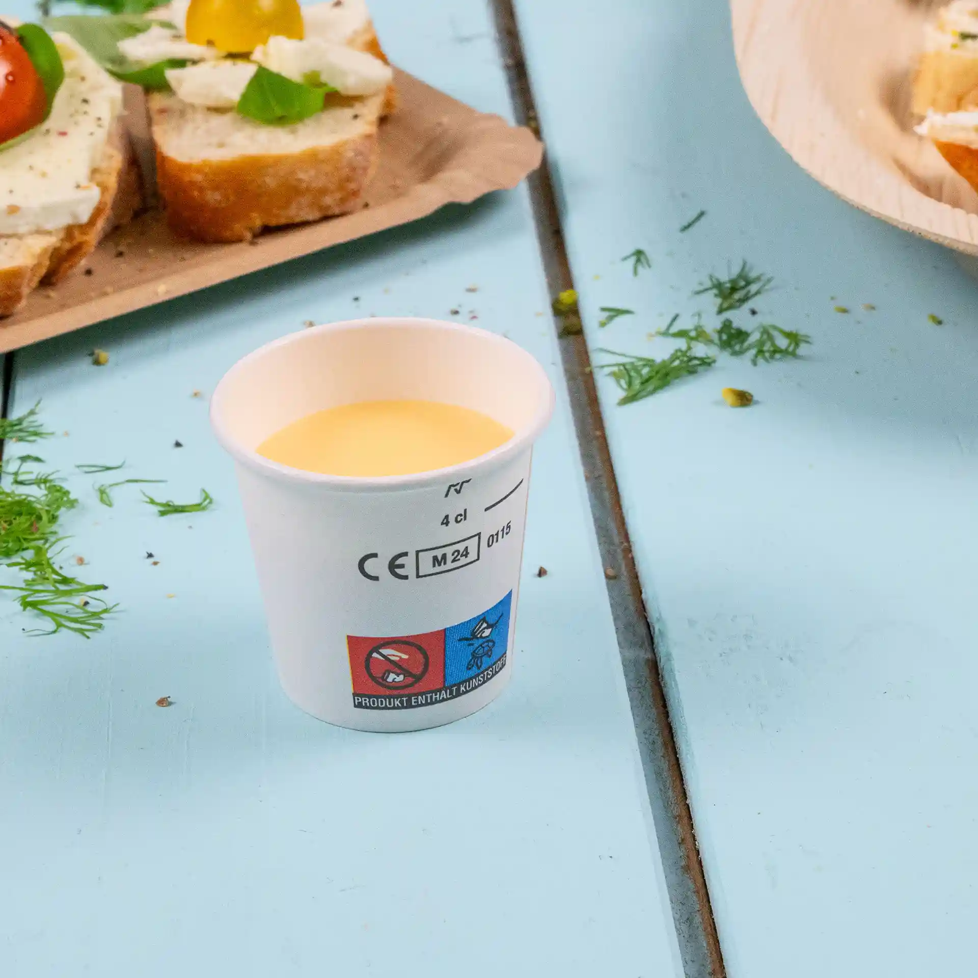Witte beker gevuld met gele vloeistof, geplaatst op een blauwe ondergrond met voedsel op de achtergrond.