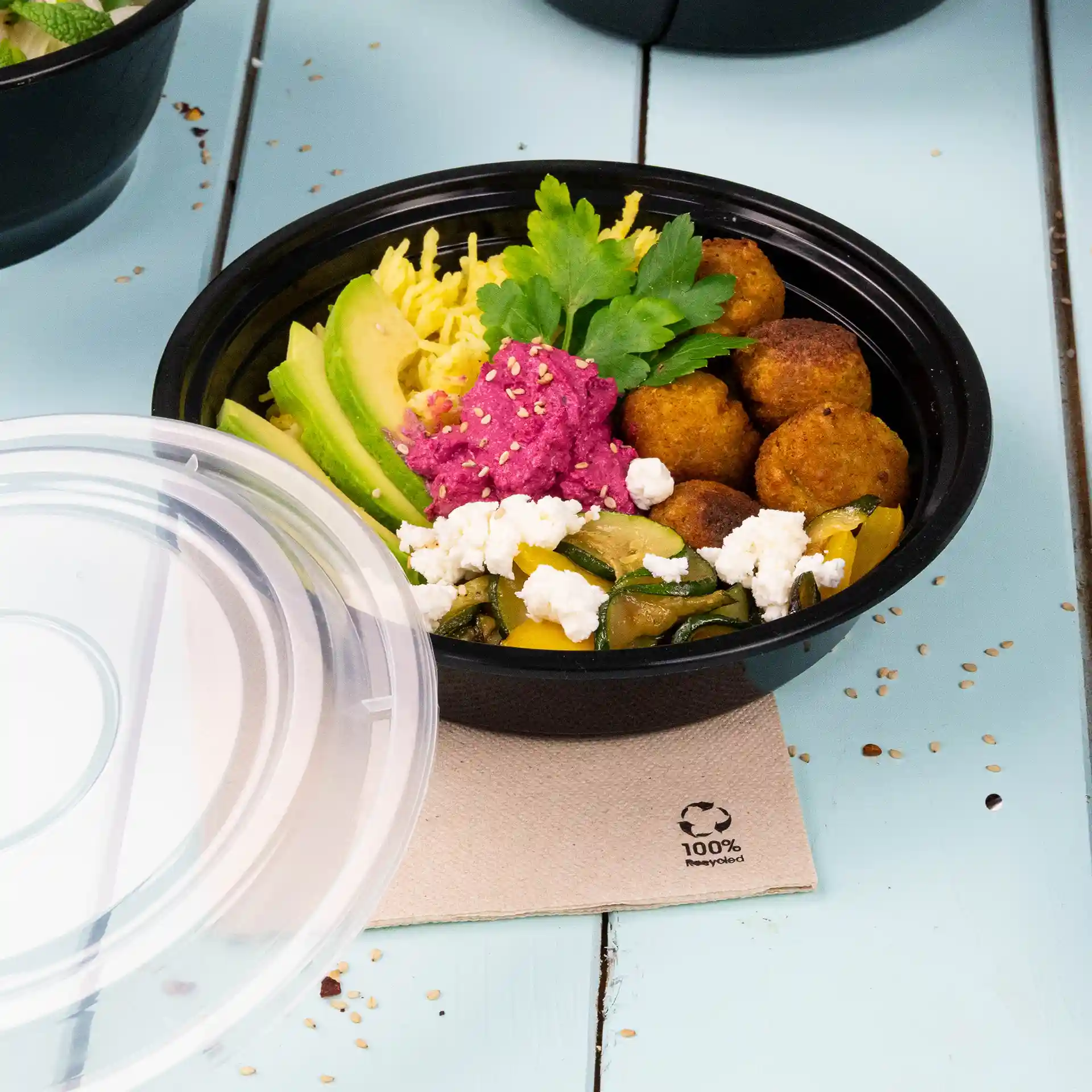 Zwarte ronde herbruikbare plaat met verschillende voedingsmiddelen, waaronder avocado, falafel en een roze spread, op een lichtblauwe ondergrond.