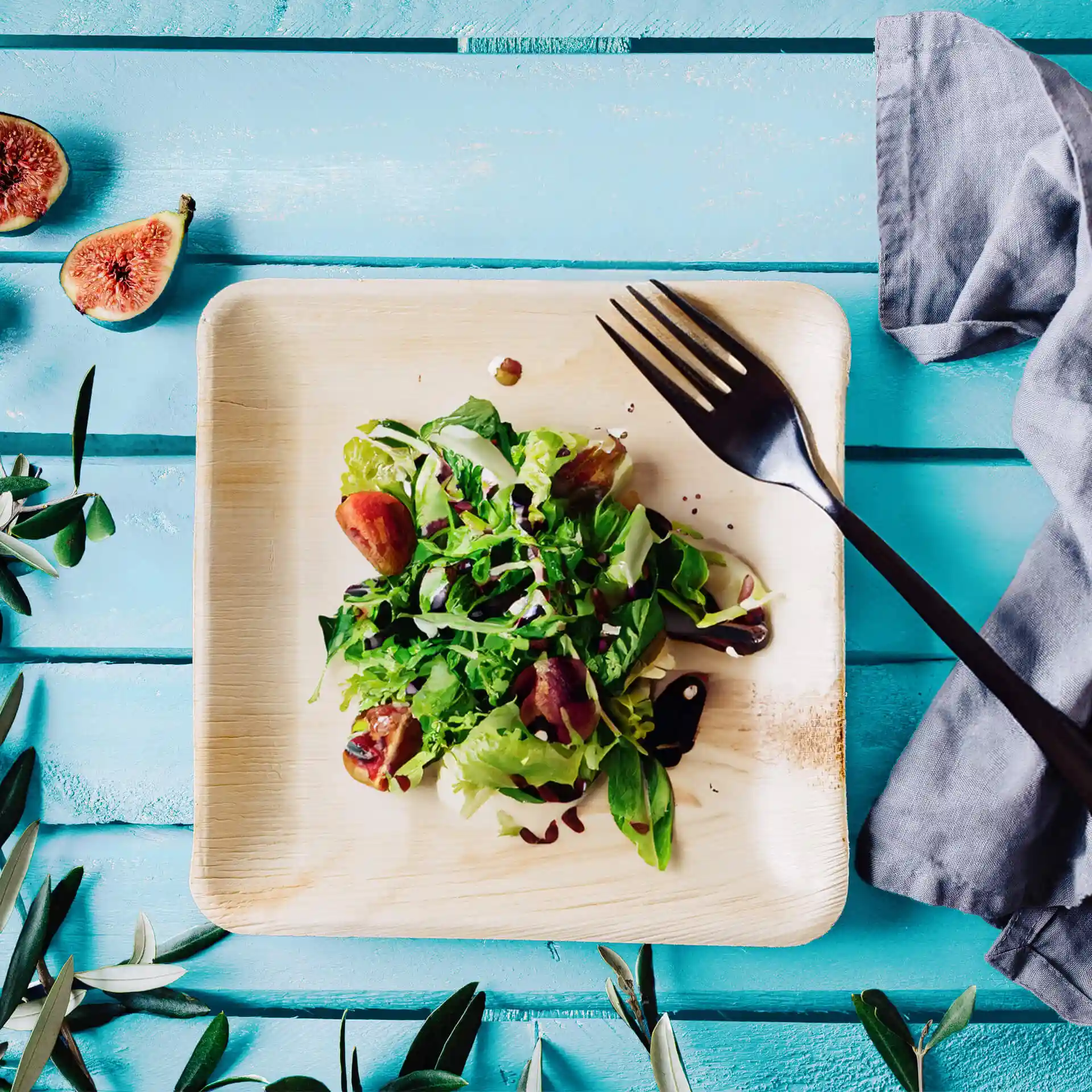 Vierkant palmblad bord met een salade, getopt met groen en vijgen, vergezeld van een zwarte vork op een blauwe houten ondergrond.